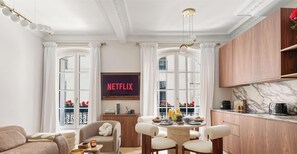 Ein Wohnzimmer mit Sofa, einem Esstisch mit Stühlen, einer Küchenzeile mit Schränken und einem großen Fenster mit Vorhängen.