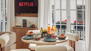 Ein Frühstückstisch mit Tellern, Früchten und einer Saftkaraffe, ein Bücherregal mit Büchern und ein Schild mit der Aufschrift 'NETFLIX'.