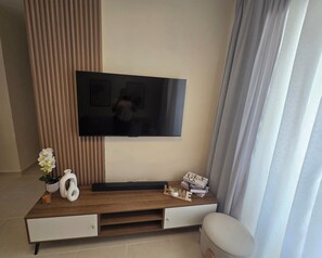 Ein modernes Wohnzimmer mit einem flachbildfernseher, der an einer Holzkonsole befestigt ist, einem kleinen weißen Hocker und einer Topfpflanze.