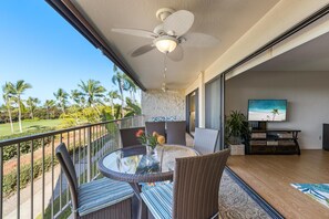Ein Balkon mit rundem Tisch, Korbstühlen, einem Deckenventilator und einem Fernseher auf einem Standfuß.