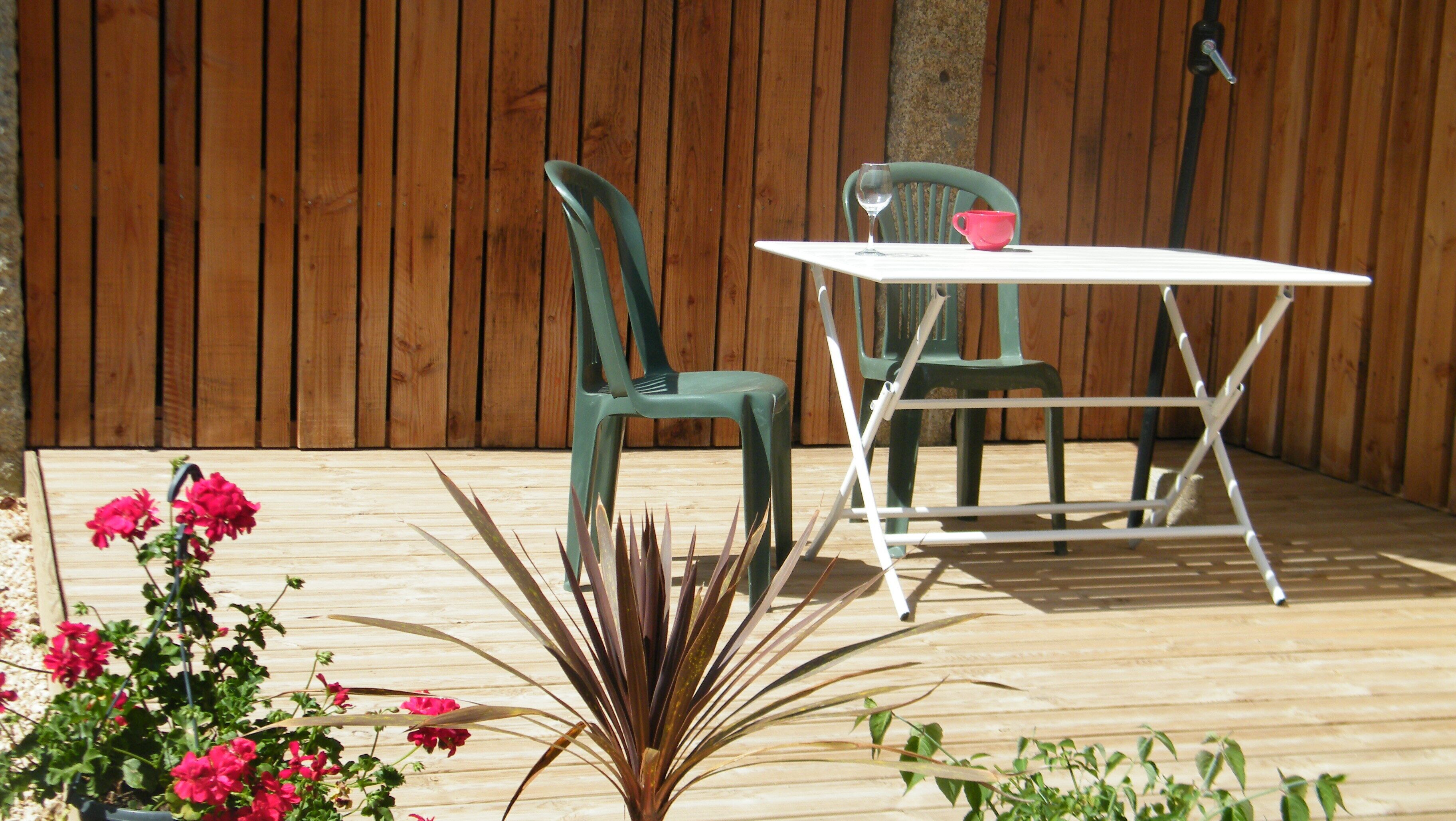 Une terrasse extérieure avec une plateforme en bois, deux chaises vertes, une table blanche et un pot de fleurs contenant des fleurs roses.