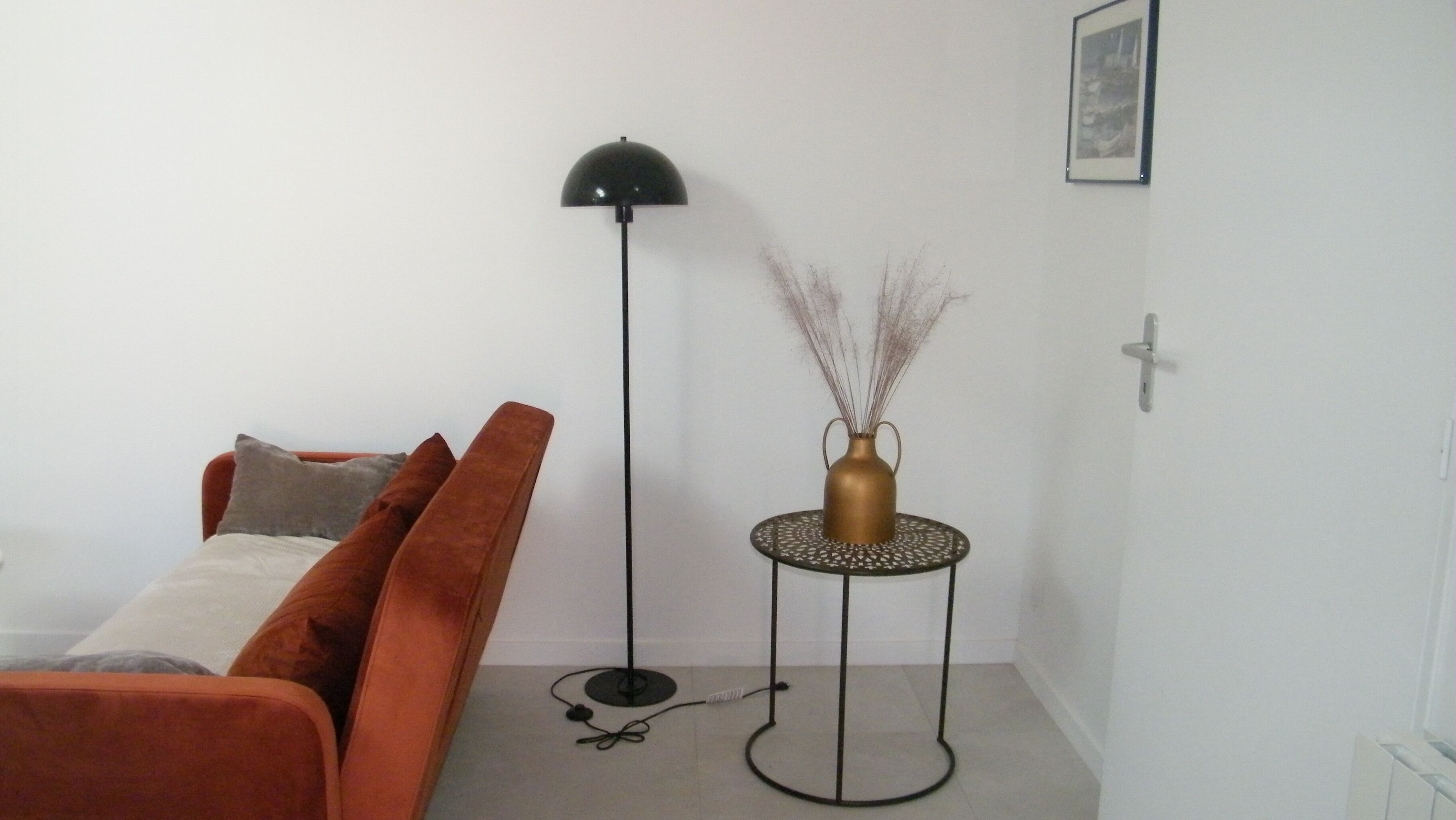 A living room with a sofa, a floor lamp, a small table with a vase, and a framed picture on the wall.