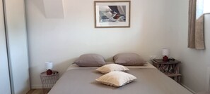 Ett sovrum med en säng, kuddar, ett sängbord med en lampa, en inramad tavla på väggen och ett fönster med gardiner.