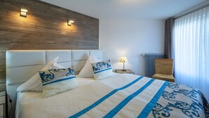 Une chambre d’hôtel avec un grand lit, deux lampes fixées au mur, une table de chevet avec une lampe, un fauteuil en osier et une fenêtre avec des rideaux.