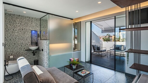 Una habitación moderna con zona de spa, pared de vidrio que da a un área de estar exterior y un paisaje urbano visible.