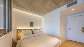 A modern bedroom with a large bed, a bedside table, a headboard with built-in lighting, a door, and a window.