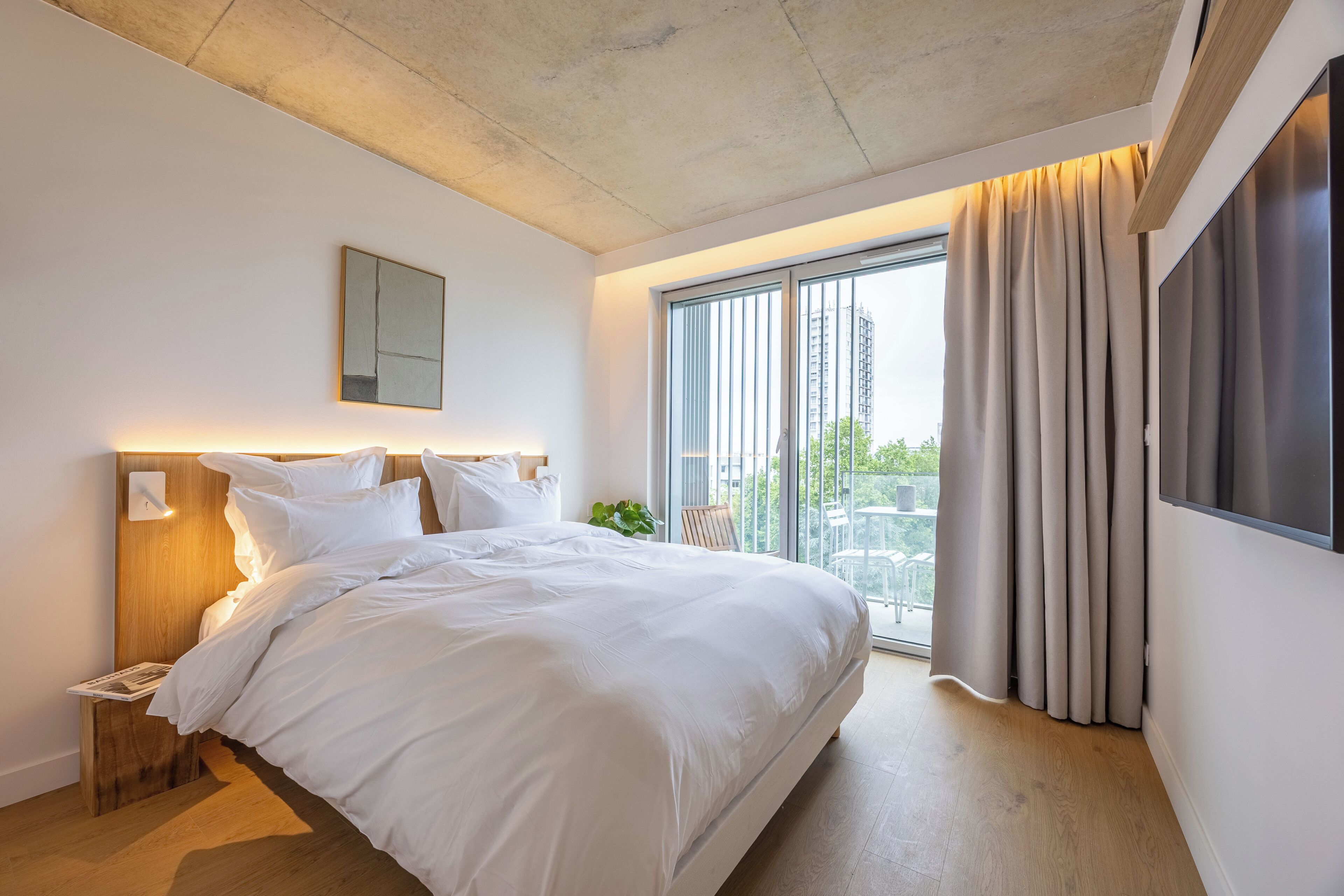 Een moderne hotelkamer met een groot bed, een nachtkastje, een tv aan de muur en een balkon met gordijnen.