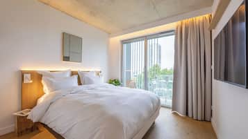 Una habitación de hotel moderna con una cama grande, una mesita de noche, un televisor montado en la pared y un balcón con cortinas.