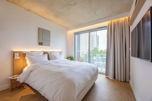 Una habitación de hotel moderna con una cama grande, una mesita de noche, un televisor montado en la pared y un balcón con cortinas.