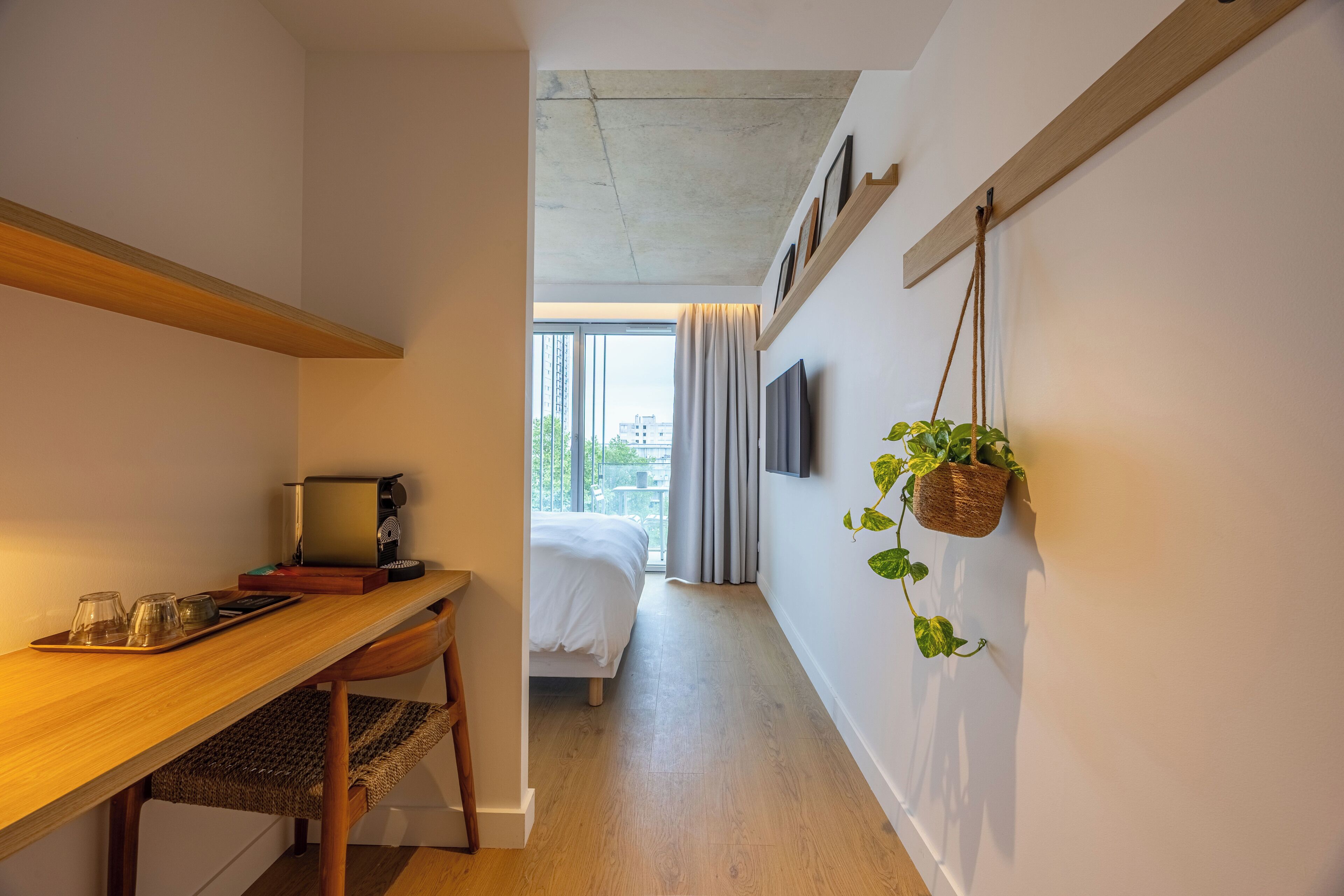 Habitación de hotel compacta con escritorio de madera, cama, televisor montado en la pared y un ventanal con cortinas.