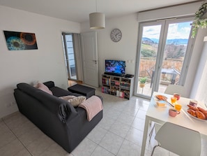 Ein Wohnzimmer mit schwarzem Sofa, Fernseher, Bücherregal und einem Essbereich mit Frühstücksangebot.