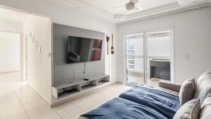 A modern living room with a flat-screen TV, a ceiling fan, and a sliding glass door leading to a balcony. - Beautiful beachfront apartment with unobstructed ocean views, pool, and sauna. (Bertioga)