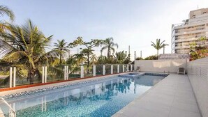 A swimming pool with a tiled edge, surrounded by palm trees and a building. - Beautiful beachfront apartment with unobstructed ocean views, pool, and sauna. (Bertioga)