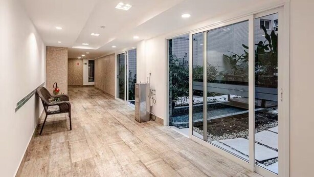 A corridor with a chair, a vending machine, and large sliding glass doors leading to an outdoor area with plants and a bench. - Beautiful beachfront apartment with unobstructed ocean views, pool, and sauna. (Bertioga)
