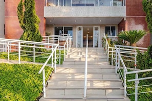 Un edificio con una escalera que lleva a una puerta, rodeado de follaje y una barandilla metálica.