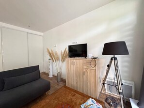 A living room with a sofa, a TV on a wooden cabinet, a lamp, and a radiator. - Charming house, pets welcome - 4 guests (Saint-Jean-de-Monts)