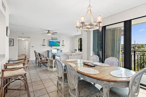A dining area with a wooden table, chairs, and a chandelier. There is a television on the wall, a ceiling fan, and a view of the outdoors through large windows. - ⭐Enclave 203B ☀Walk to the Beach ☀Community Pool~Tennis and Volleyball Courts⭐ (Destin)