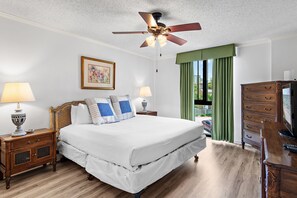 Una habitación de hotel con una cama, mesitas de noche, cómoda y un ventilador de techo, además de un ventanal con cortinas.