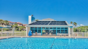 Una piscina con zona cubierta, un edificio con grandes ventanales y un cielo azul despejado.