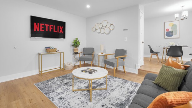 Ein modernes Wohnzimmer mit einem Flachbildfernseher, grauen Sofas, einem Couchtisch und einem Essbereich mit Tisch und Stühlen.