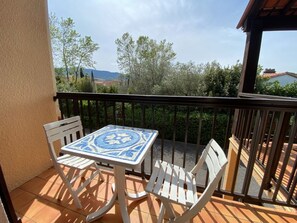Ein Balkon mit einem gefliesten Tisch und zwei weißen Stühlen, mit Blick auf eine Landschaft aus Bäumen und Gebäuden.