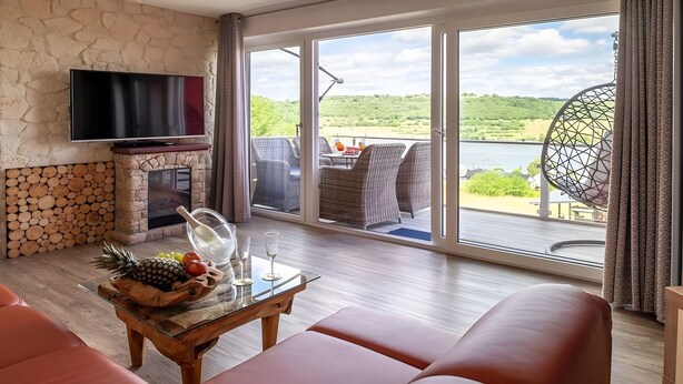 A room with a stone fireplace, a flat-screen TV, wicker furniture, a glass coffee table with fruits, and a view of a lake and greenery. - Exclusive Apartment for 4 People (56 m²) in Schalkenmehren with Lake View (Schalkenmehren)
