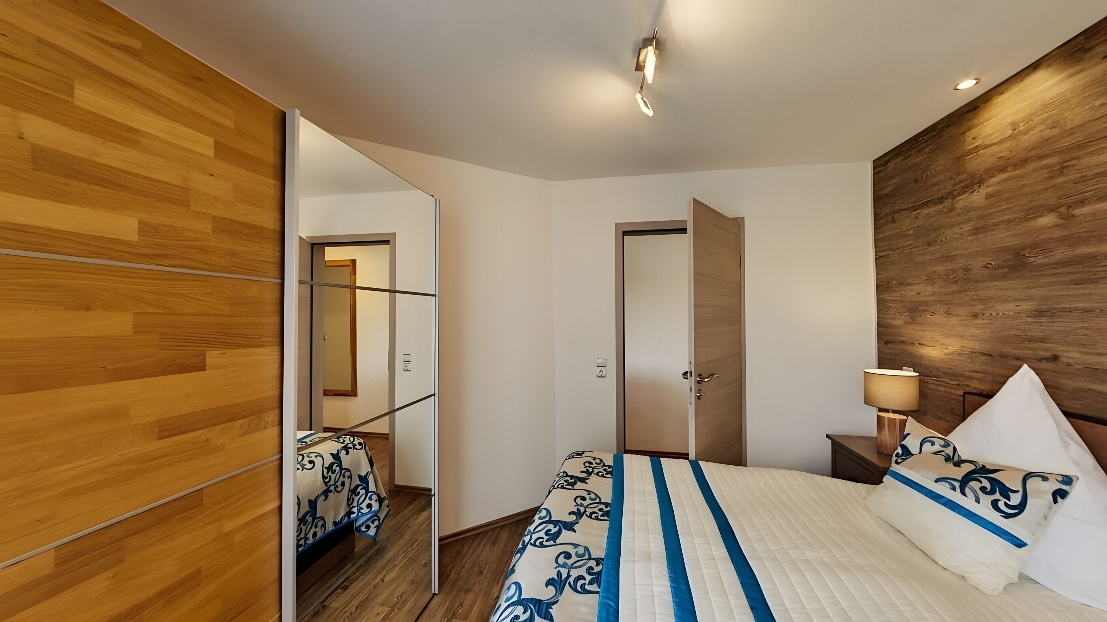 A modern bedroom with a wooden floor, a bed with a patterned bedspread, a bedside lamp, and a mirror.