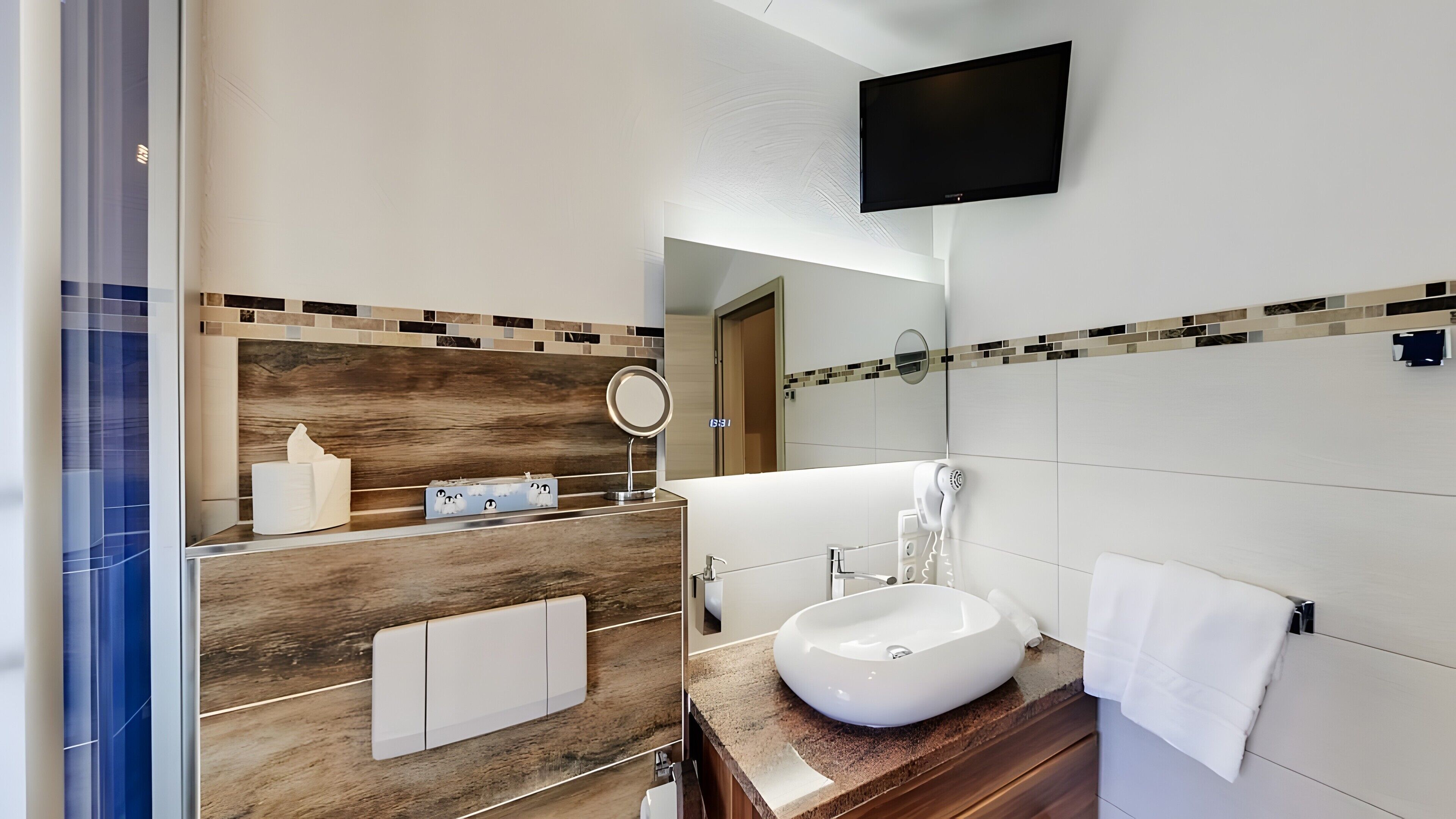 Un baño moderno con tocador de madera, un espejo grande, televisor montado en la pared y un lavamanos blanco.