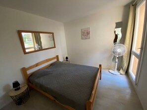 Ein Schlafzimmer mit einem hölzernen Bett, einem Nachttisch mit Lampe, einem Standventilator und einem Fenster mit Vorhängen.