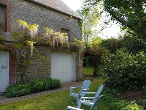 Ein Steinhaus mit einer weißen Garagentür, einem Garten mit grünen Sträuchern und zwei blauen Adirondack-Stühlen.