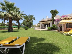 Ein gepflegter Rasen mit zwei gelben Liegestühlen, eine Terrasse mit Tisch und Stühlen sowie ein Gebäude mit Ziegeldach und Balkon.