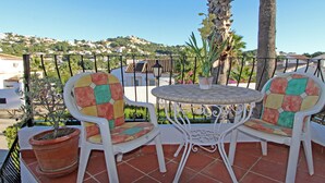 Un patio con dos sillas y una mesa, rodeado por una cerca de metal, con vistas a casas y vegetación al fondo.