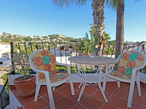 A patio with two chairs and a table, surrounded by a metal fence, with a view of houses and greenery in the background. - Cozy Moraira Apartment with Shared Pool, Garden, and Close to Beach (Moraira)