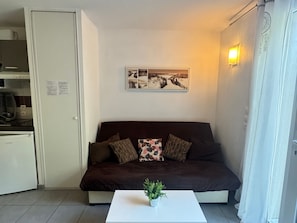 A compact living space with a sofa, a small coffee table, a wall-mounted picture, and a kitchen area. - Country house - 2 guests (Auberville)