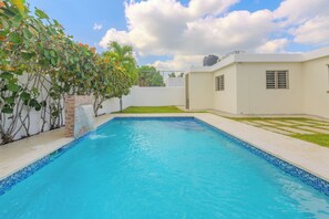 Piscina retangular com azulejos azuis, rodeada por um edifício branco e uma pequena área gramada.
