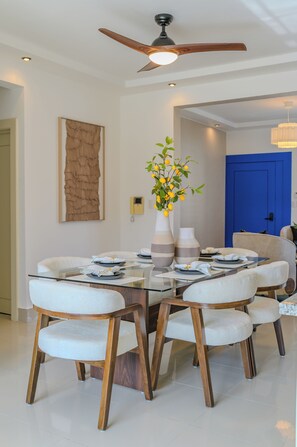 Uma sala de jantar com uma mesa de vidro, cadeiras estofadas brancas, um ventilador de teto e uma porta azul ao fundo.