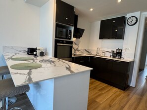 A modern kitchen with a marble countertop, black cabinets, a microwave, oven, and a clock on the wall. - Renovated apartment, lake view, private parking (Aix-les-Bains)