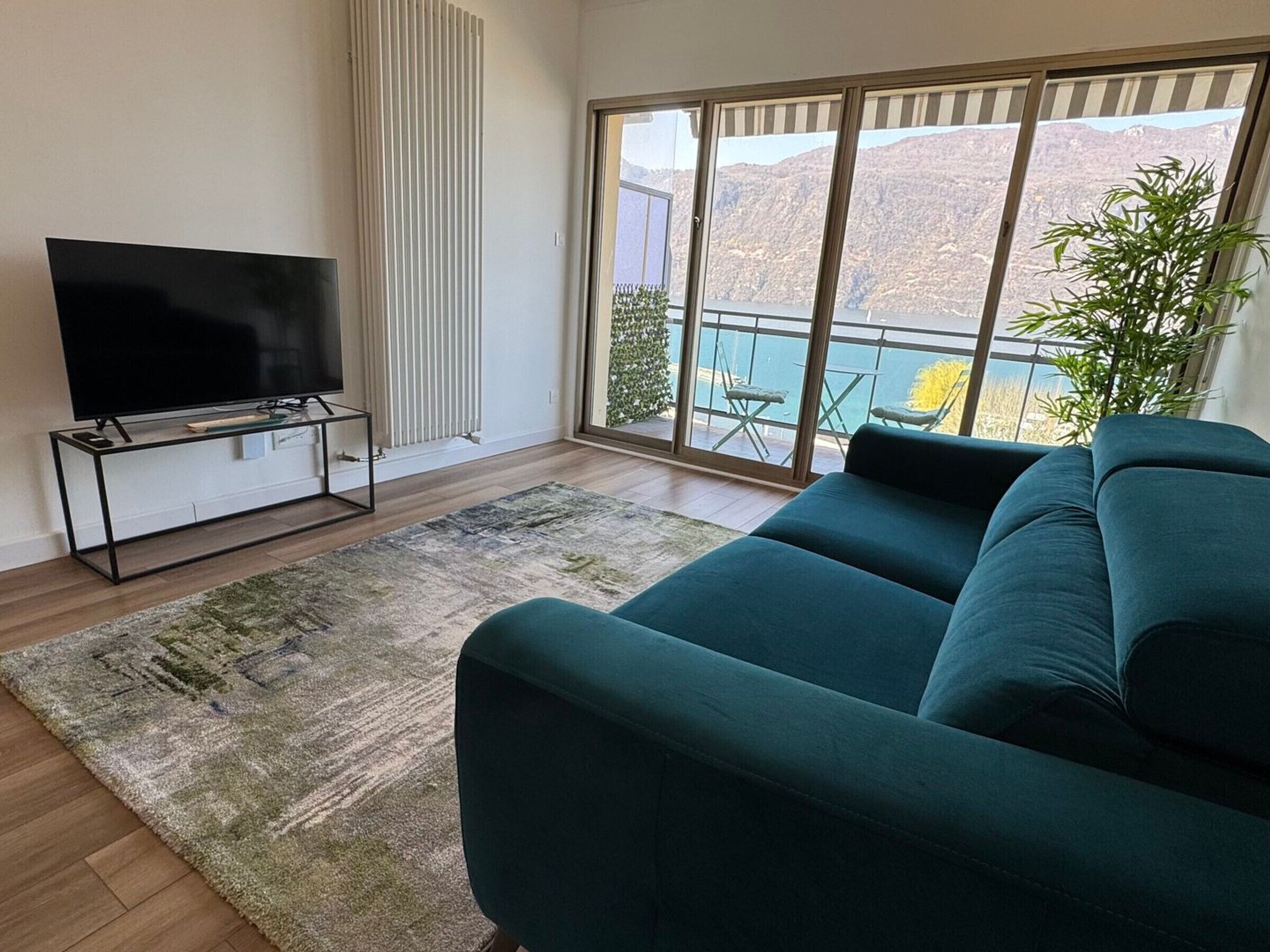 A living room with a large TV on a stand, a teal sofa, a patterned rug, and a balcony with a view of mountains.