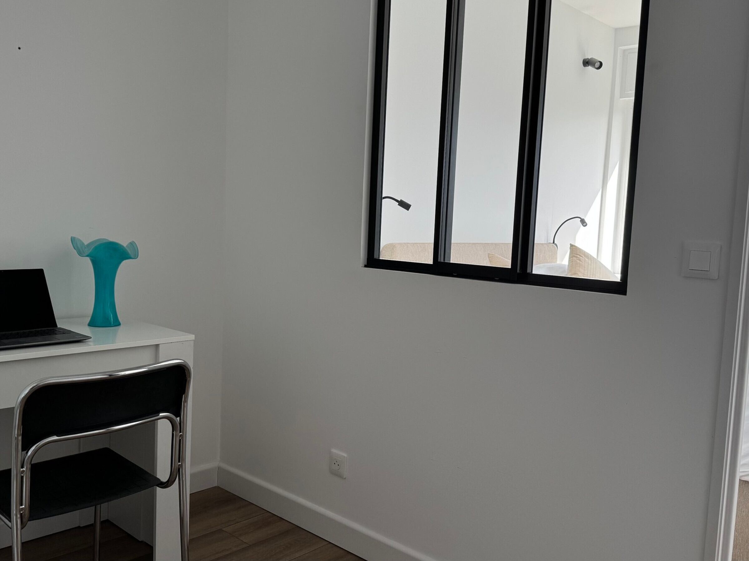 Una habitación pequeña con un escritorio blanco, una silla, una computadora portátil, un florero azul turquesa y una ventana que da a un baño.