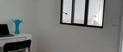 Una habitación pequeña con un escritorio blanco, una silla, una computadora portátil, un florero azul turquesa y una ventana que da a un baño.