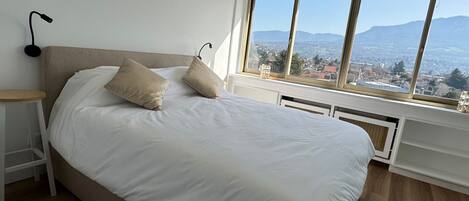 Un dormitorio con cama, almohadas, mesita de noche, ventana con vistas a montañas y una lámpara de pared.