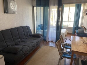 Ein Wohnzimmer mit einem dunklen Sofa, einem Esstisch, Korbstühlen und Blick durch eine Glastür.