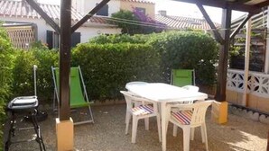 Une terrasse couverte avec une table et des chaises, entourée de verdure et avec un bâtiment en arrière-plan.
