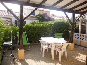 Eine überdachte Terrasse mit Tisch und Stühlen, umgeben von viel Grün und einem Gebäude im Hintergrund.