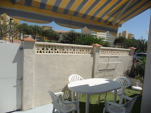 Eine Terrasse mit einem weißen Tisch und Stühlen, einem gestreiften Sonnensegel und einem dekorativen Zaun.