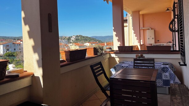 A balcony with a table and chairs, potted plants, and a view of buildings and hills.