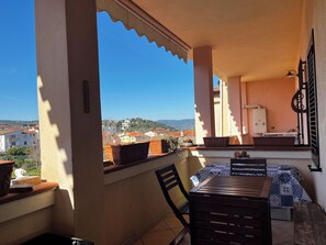 Varanda com mesa e cadeiras, plantas em vasos e vista para edifícios e colinas.