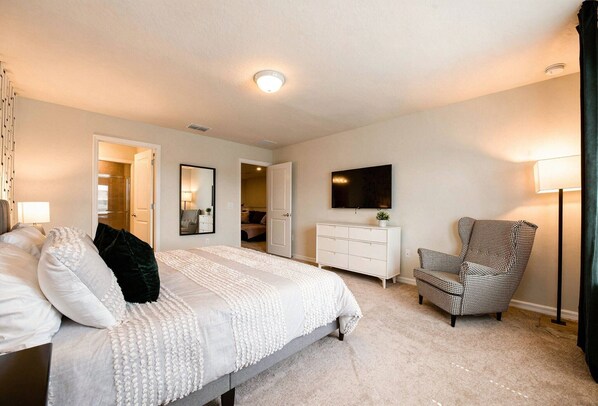 Un dormitorio con cama, cómoda, silla, televisor y una puerta que da a otra habitación.