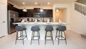 A modern kitchen with dark cabinets, stainless steel appliances, a marble island with bar stools, and pendant lighting. - Windsor Island 5BR Home Near Disney (Davenport)