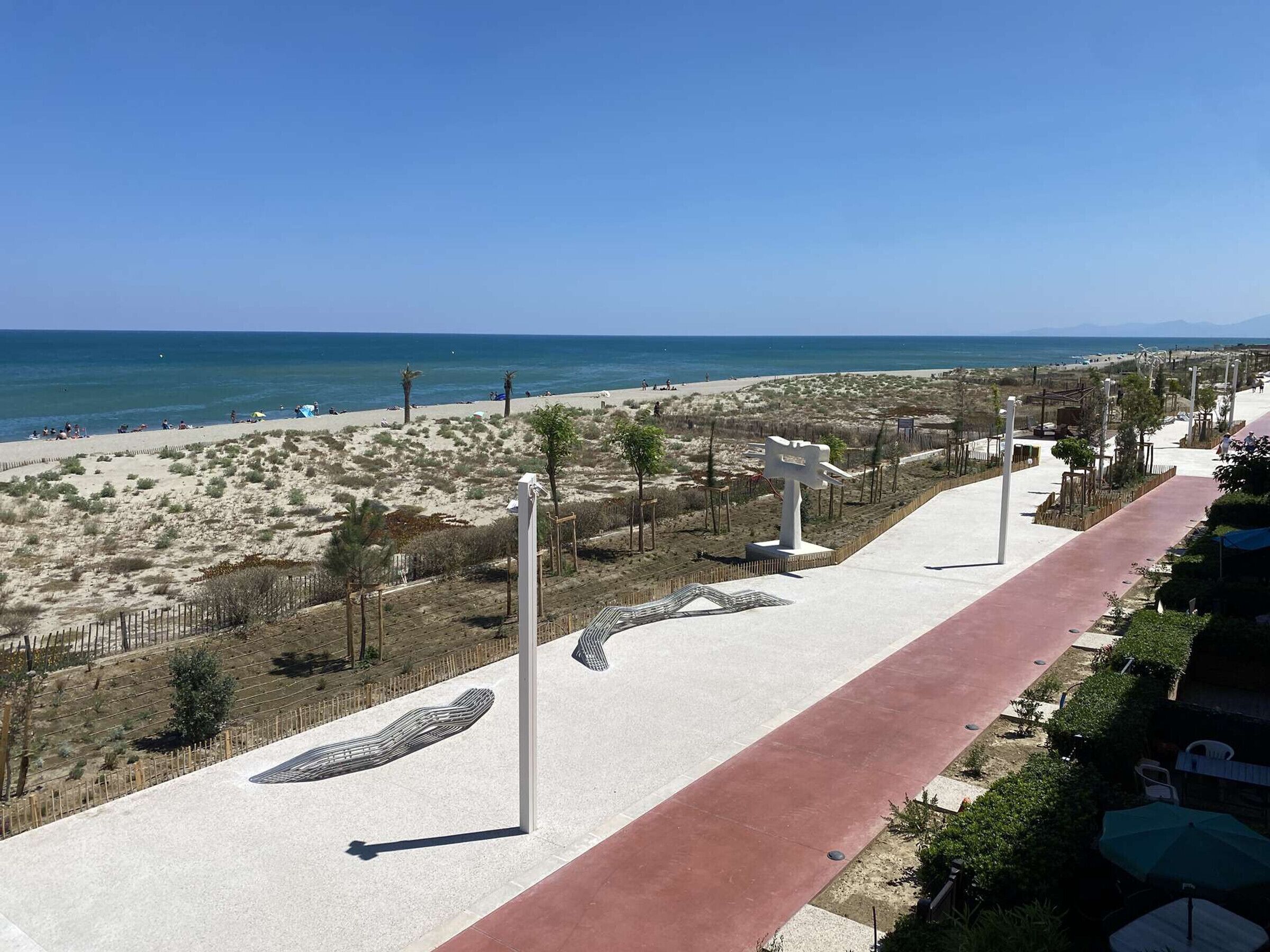 A coastal walkway with a red path, white sculptures, and a beach in the background.